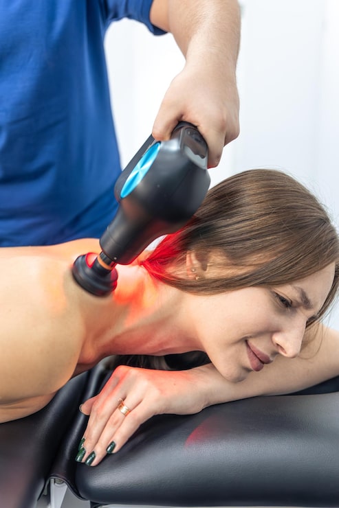 Professional photograph of a red and near-infrared laser therapy device positioned over a patient's joint or shoulder — warm red glow on the skin, calm clinical setting with sage and warm cream tones