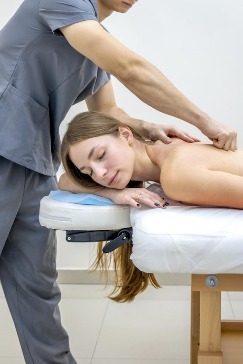 Professional photograph of a therapist's hands performing gentle lymphatic drainage strokes along a patient's neck and collarbone area — softly lit treatment room with calm spa-like atmosphere, sage and warm cream tones