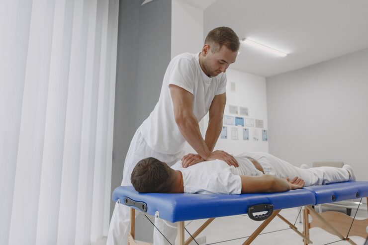 Photograph of a patient on a treatment table while a practitioner performs targeted muscle release on the upper back — focused therapy with sage-toned linens, warm natural lighting, calm professional atmosphere
